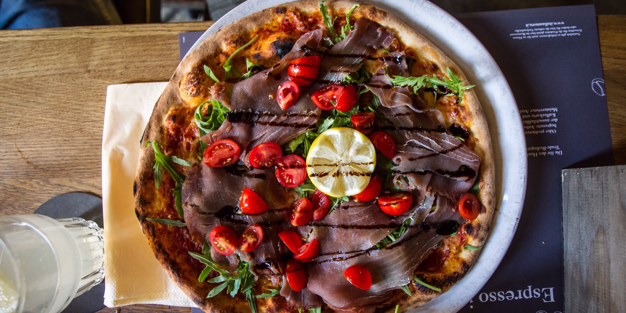 Frisch gebackene Pizza mit luftgetrocknetem Schinken, Rucola, Kirschtomaten und gegrillter Zitrone in einer Innsbrucker Pizzeria.