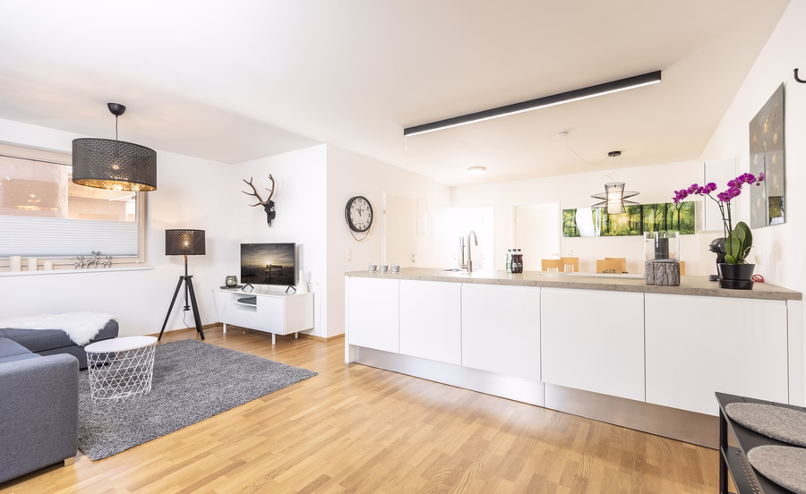 Heller offener Wohnbereich mit großer weißer Kücheninsel, grauem Sofa, Fernsehecke und modernen schwarzen Leuchten.