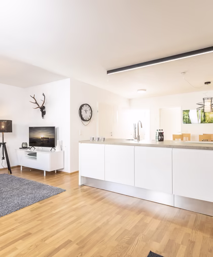 Heller offener Wohnbereich mit großer weißer Kücheninsel, grauem Sofa, Fernsehecke und modernen schwarzen Leuchten.