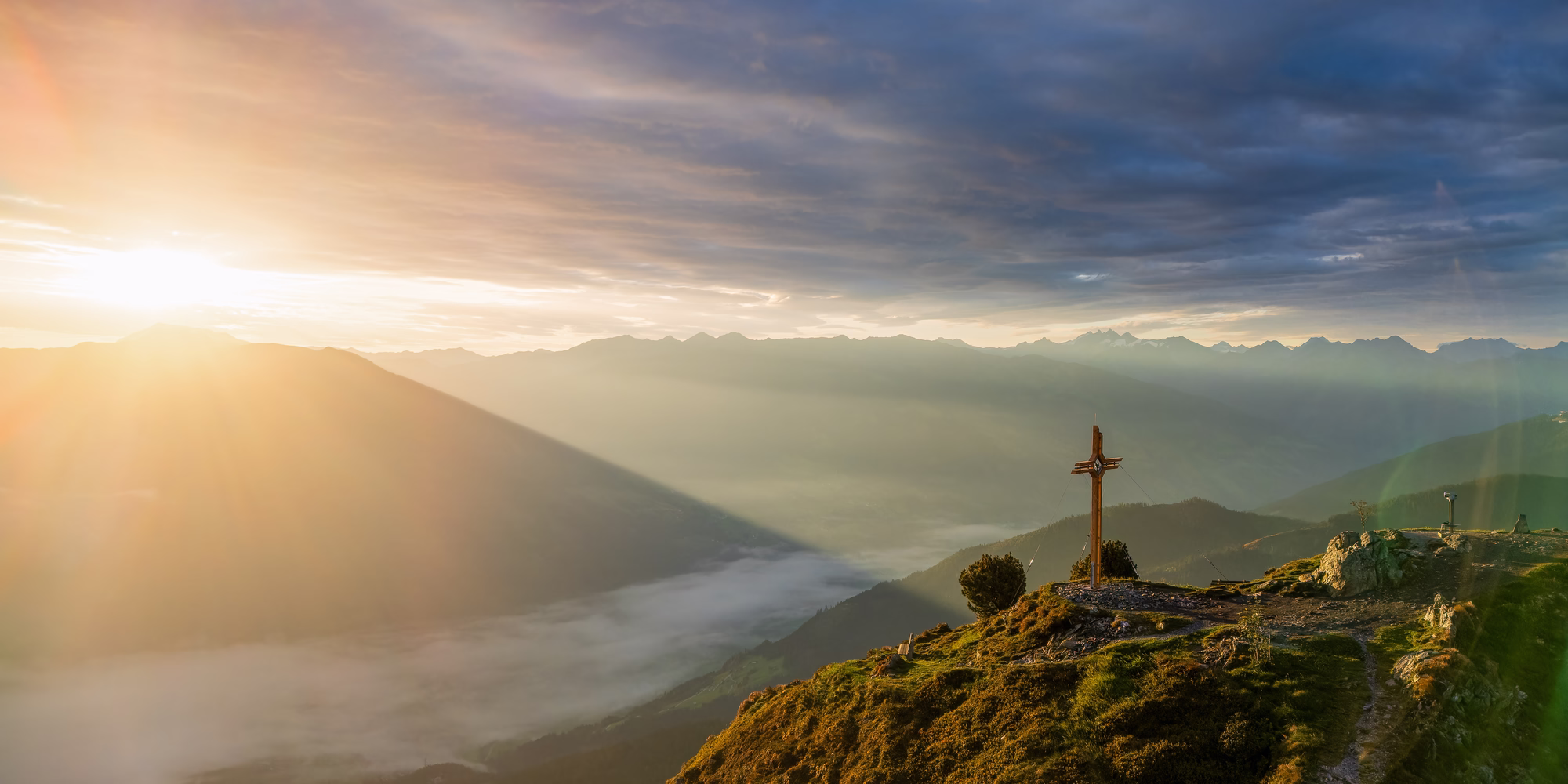 Gipfelkreuz am Spieljoch im goldenen Morgenlicht mit Blick über das nebelbedeckte Zillertal und die umliegenden Bergketten.