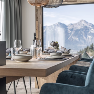 Einladend gedeckter Holztisch mit blauen Sesseln und großer Fensterfront, die den Blick auf das eindrucksvolle Bergpanorama freigibt.