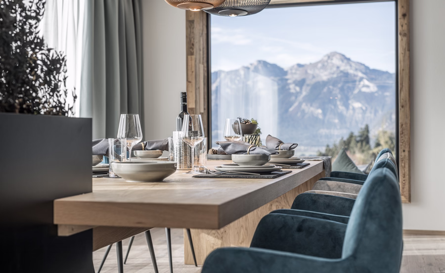 Einladend gedeckter Holztisch mit blauen Sesseln und großer Fensterfront, die den Blick auf das eindrucksvolle Bergpanorama freigibt.