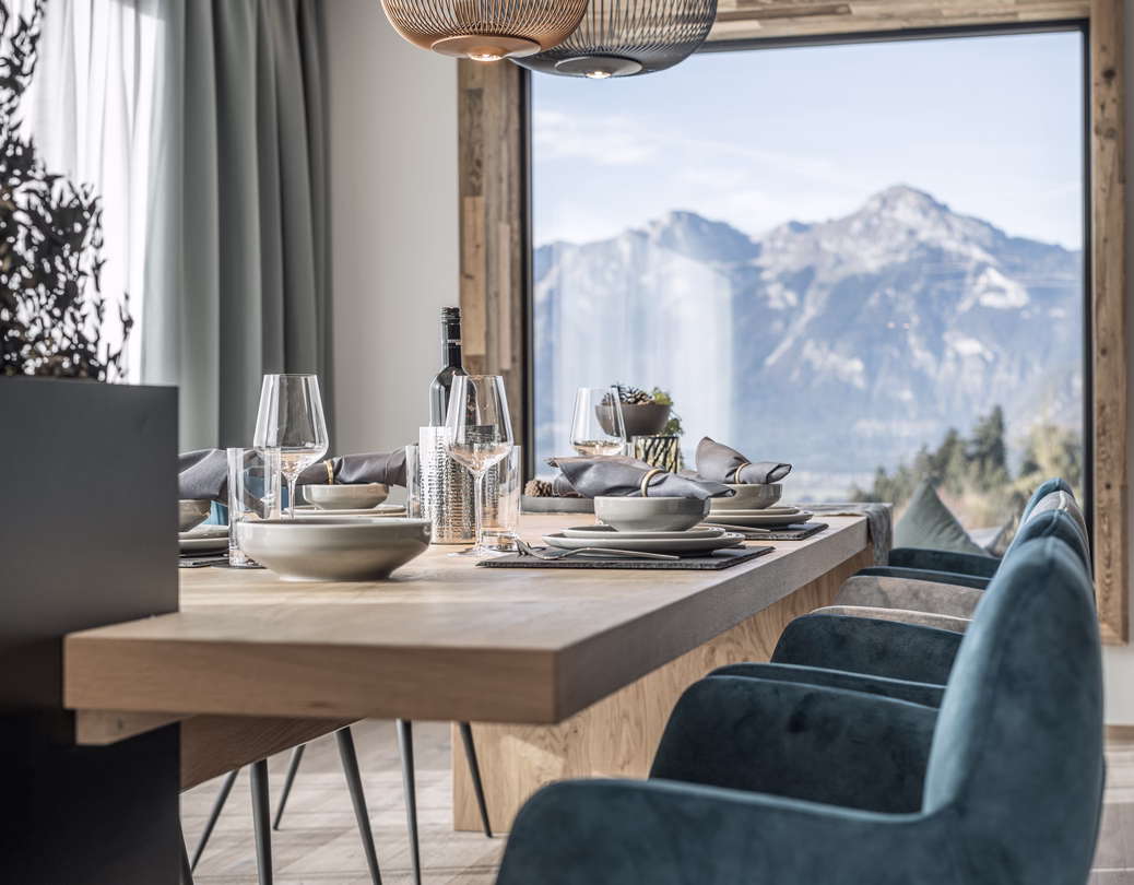 Einladend gedeckter Holztisch mit blauen Sesseln und großer Fensterfront, die den Blick auf das eindrucksvolle Bergpanorama freigibt.