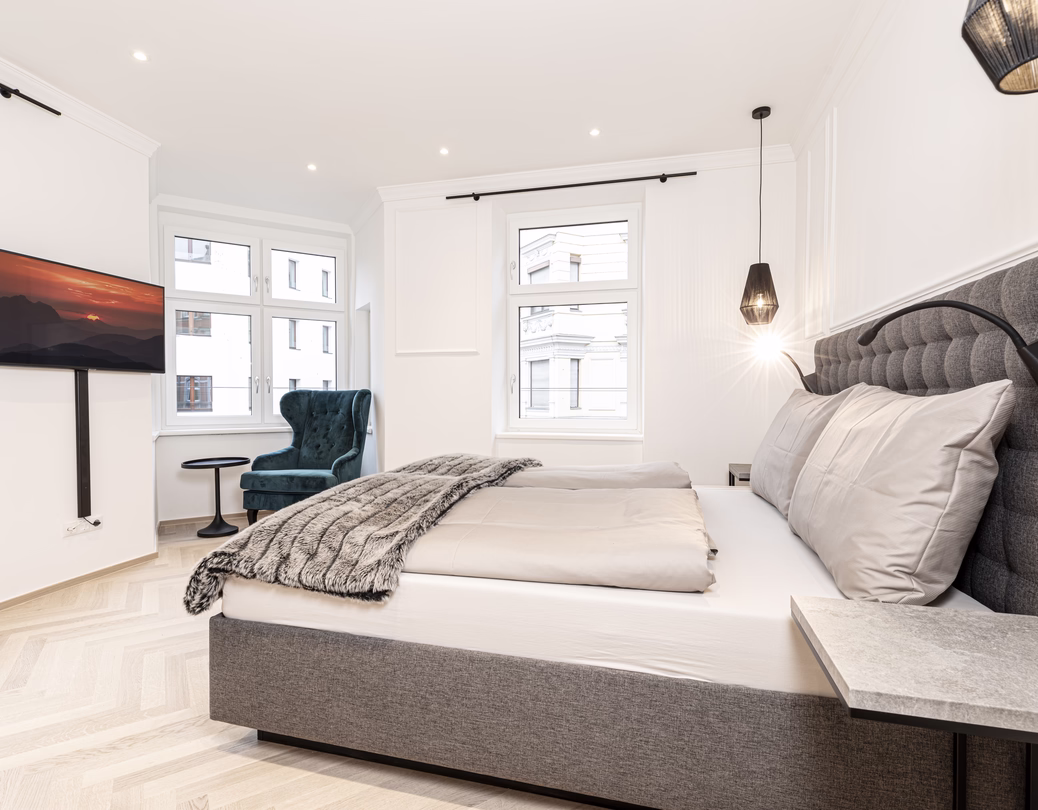 Komfortables Schlafzimmer mit großem Doppelbett, gepolstertem Kopfteil, Fernseher und türkisfarbenem Ohrensessel.