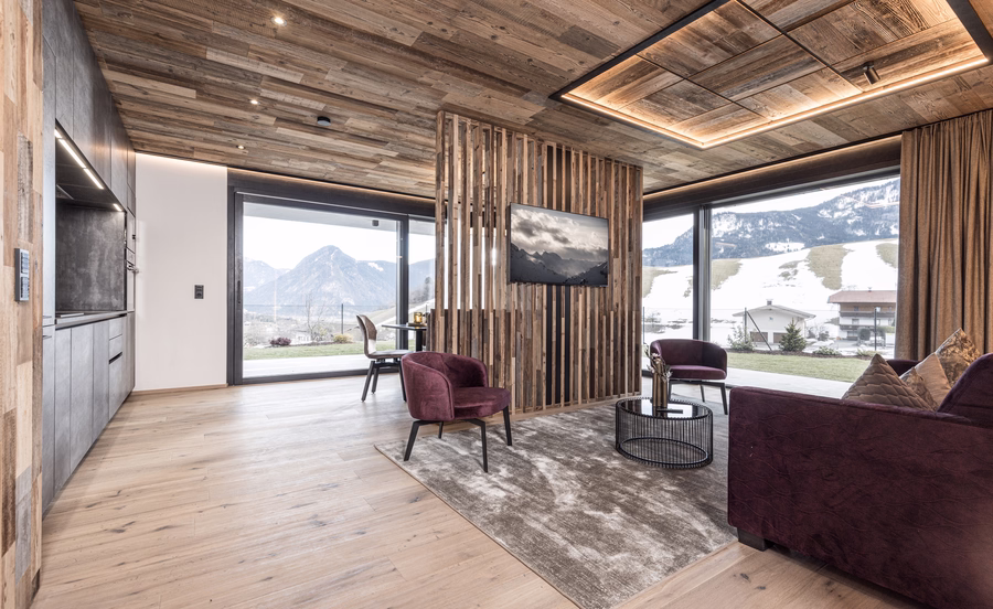 Modernes Wohnzimmer mit burgunderfarbenen Sitzmöbeln, Fernseher, rustikaler Holzdecke und großen Panoramafenstern zur Winterlandschaft.