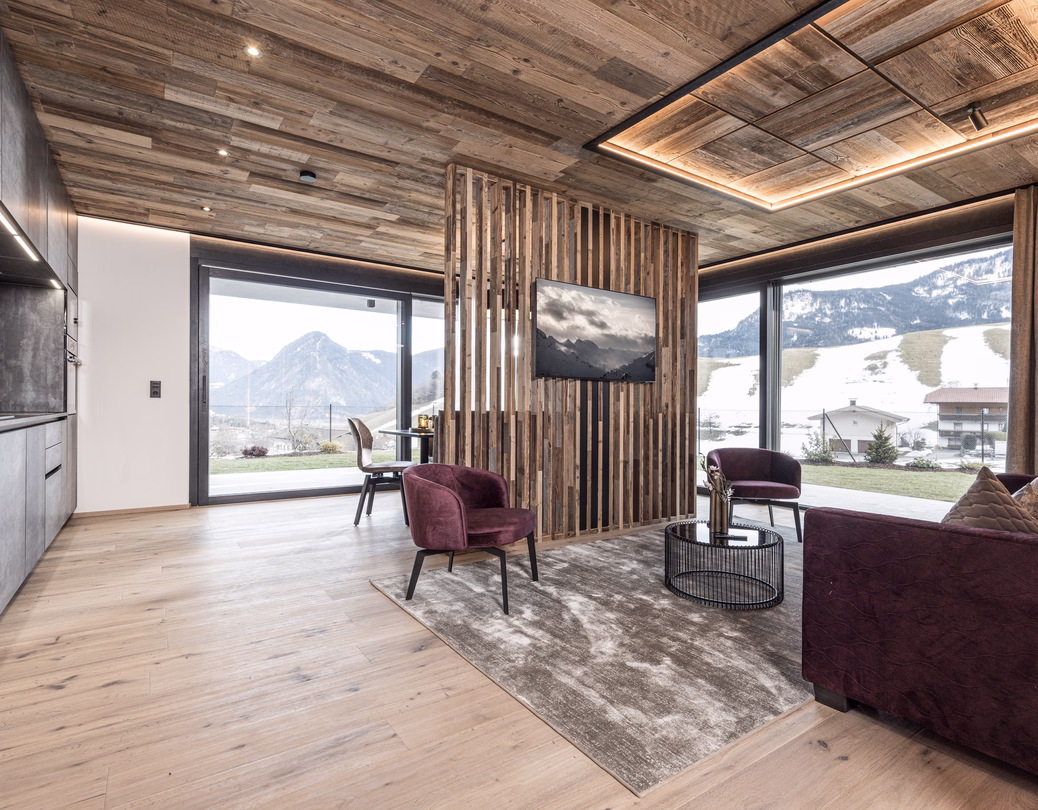 Modernes Wohnzimmer mit burgunderfarbenen Sitzmöbeln, Fernseher, rustikaler Holzdecke und großen Panoramafenstern zur Winterlandschaft.