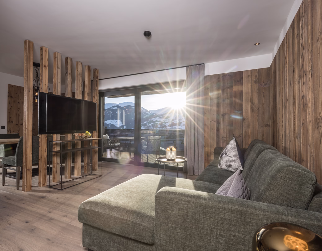 Heller Wohnbereich mit grünem Ecksofa, Fernseher und rustikalen Holzwänden sowie direktem Blick auf das sonnige Bergpanorama.