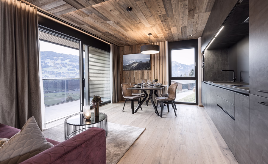 Offener Wohnbereich mit moderner Küche, gedecktem Esstisch, gemütlichem Sofa und großen Fenstern mit Blick auf die Berge.