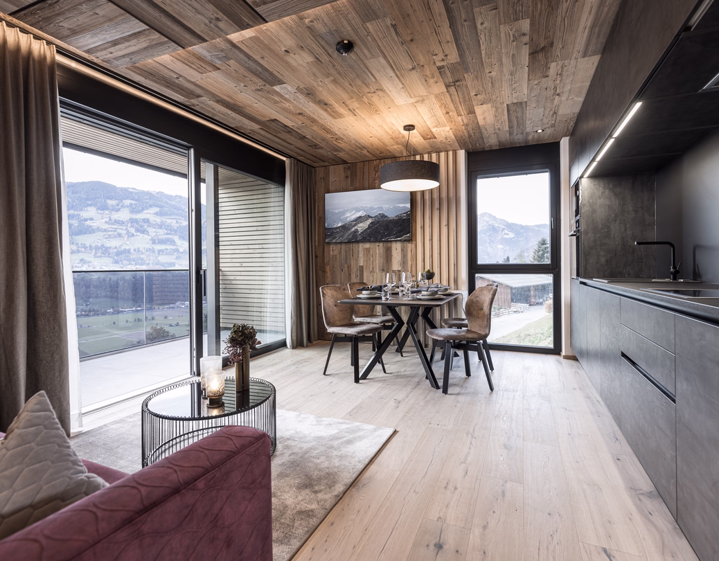 Offener Wohnbereich mit moderner Küche, gedecktem Esstisch, gemütlichem Sofa und großen Fenstern mit Blick auf die Berge.