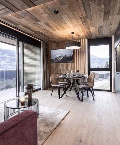 Offener Wohnbereich mit moderner Küche, gedecktem Esstisch, gemütlichem Sofa und großen Fenstern mit Blick auf die Berge.