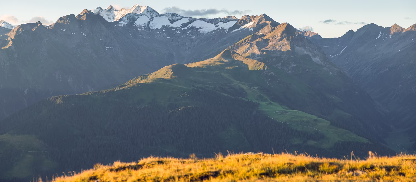 Weites Bergpanorama bei Gerlos mit sonnenbeschienenen Almwiesen, bewaldeten Hängen und schneebedeckten Gipfeln.