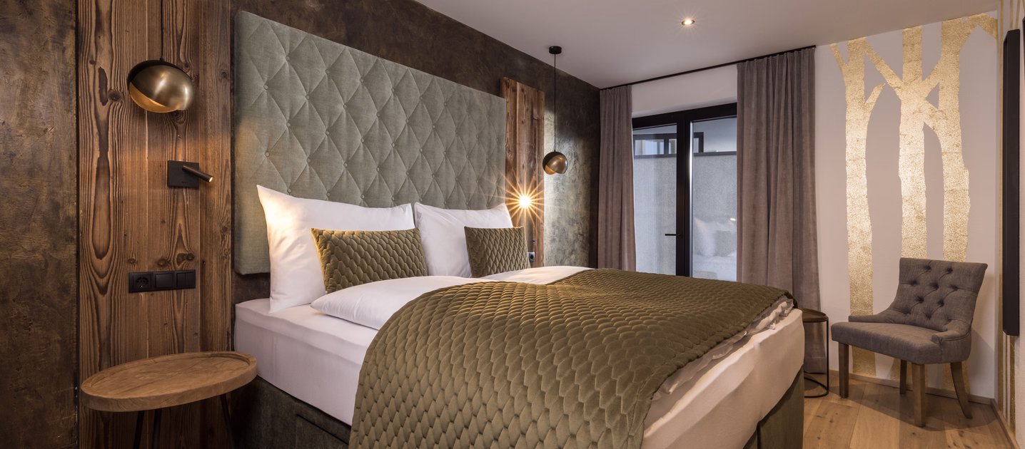 Stilvolles Doppelzimmer mit grünem Polsterbett, rustikalen Holzelementen, warmen Leuchten und goldener Baumgrafik an der Wand.
