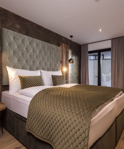 Stilvolles Doppelzimmer mit grünem Polsterbett, rustikalen Holzelementen, warmen Leuchten und goldener Baumgrafik an der Wand.