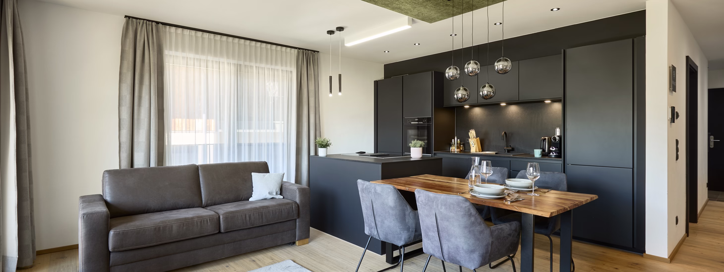Heller offener Wohn- und Essbereich mit grauem Sofa, gedecktem Holztisch und moderner dunkler Einbauküche.