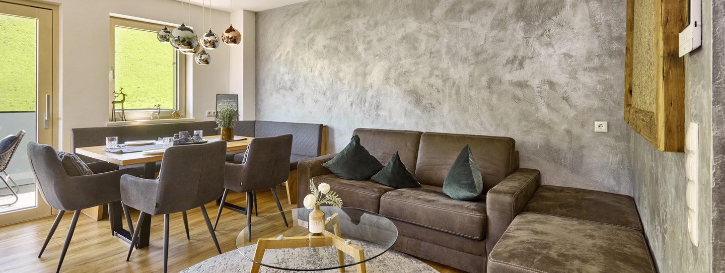 Gemütlicher Wohn- und Essbereich mit braunem Ecksofa, gedecktem Holztisch und Blick auf eine grüne Wiese.