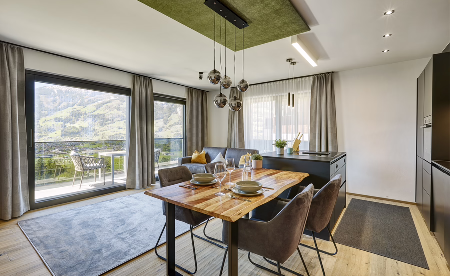Heller offener Essbereich mit gedecktem Holztisch, moderner Küche, gemütlichem Sofa und großzügigem Blick auf die Berge.