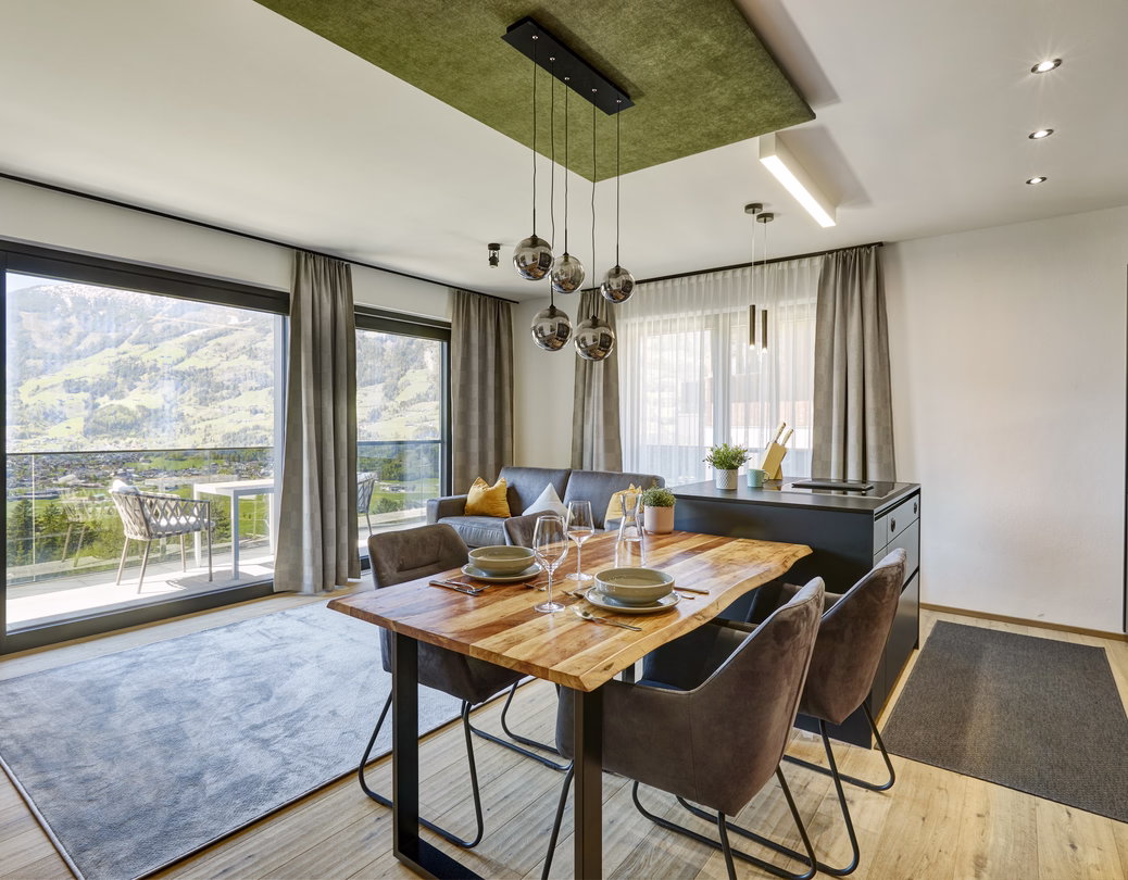 Heller offener Essbereich mit gedecktem Holztisch, moderner Küche, gemütlichem Sofa und großzügigem Blick auf die Berge.