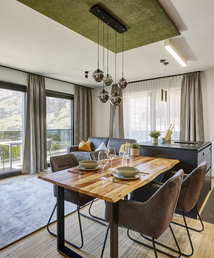 Heller offener Essbereich mit gedecktem Holztisch, moderner Küche, gemütlichem Sofa und großzügigem Blick auf die Berge.