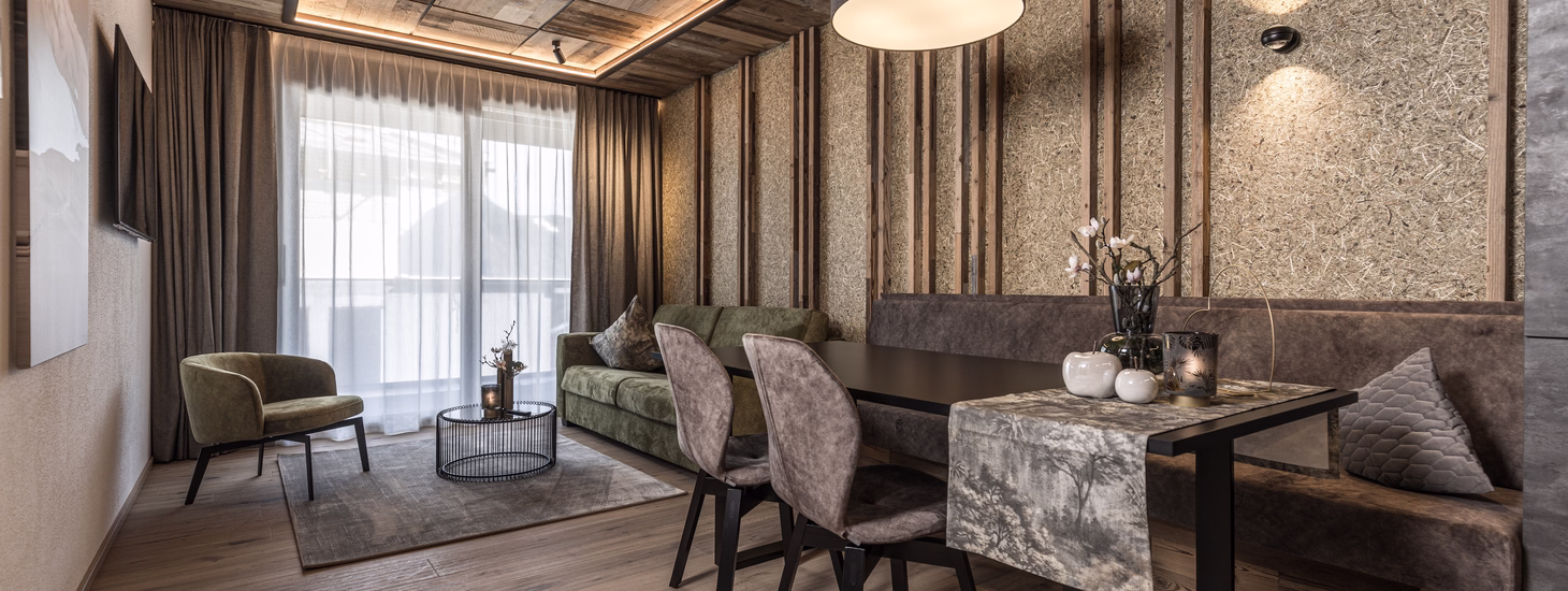 Warmer Wohn- und Essbereich mit gepolsterter Sitzbank, grünem Sofa, Holzelementen und naturbelassener Wandgestaltung.