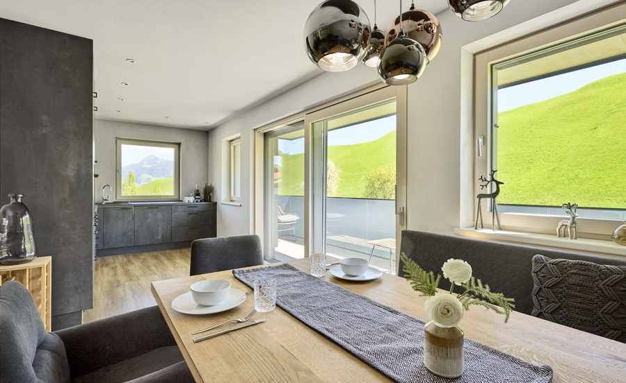 Heller Essbereich mit gedecktem Holztisch, moderner Küche, Balkonzugang und weitem Blick auf grüne Wiesen und Berge.