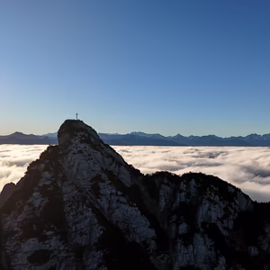 Felsiger Gipfel des Ebner Jochs mit Gipfelkreuz über einem dichten Wolkenmeer im klaren Herbstlicht.