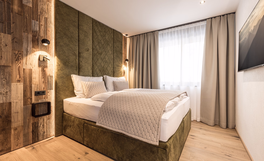 Helles Schlafzimmer mit grünem Polsterbett, hohem Kopfteil, rustikaler Holzwand, warmen Leselampen und Fernseher.