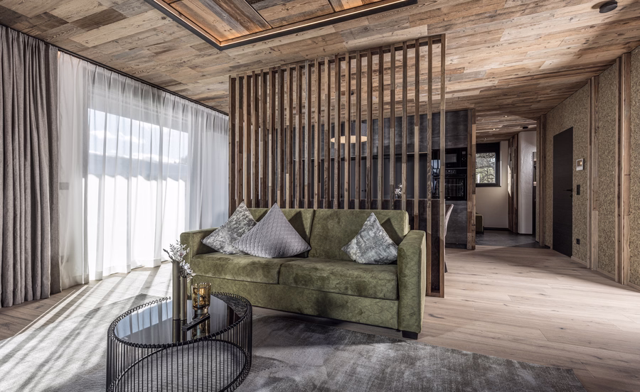 Heller Wohnbereich mit grünem Sofa, rundem Glastisch, rustikaler Holzdecke und dekorativer Raumtrennung aus Holzlamellen.