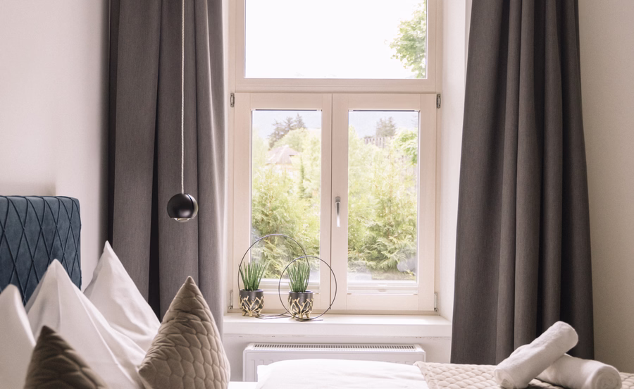 Helles Doppelbett mit grauer Tagesdecke, dekorativen Kissen und bereitgelegten Handtüchern neben einem hohen Fenster.