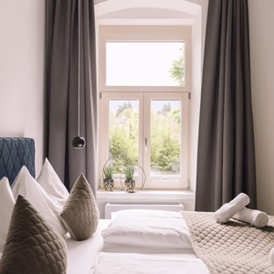 Helles Doppelbett mit grauer Tagesdecke, dekorativen Kissen und bereitgelegten Handtüchern neben einem hohen Fenster.