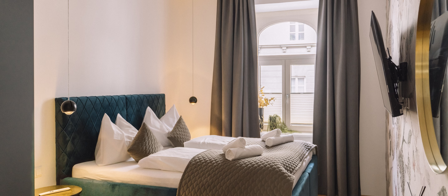 Modernes Doppelzimmer mit blauem Polsterbett, Rundbogenfenster, grauen Vorhängen, Fernseher und bereitgelegten Handtüchern.