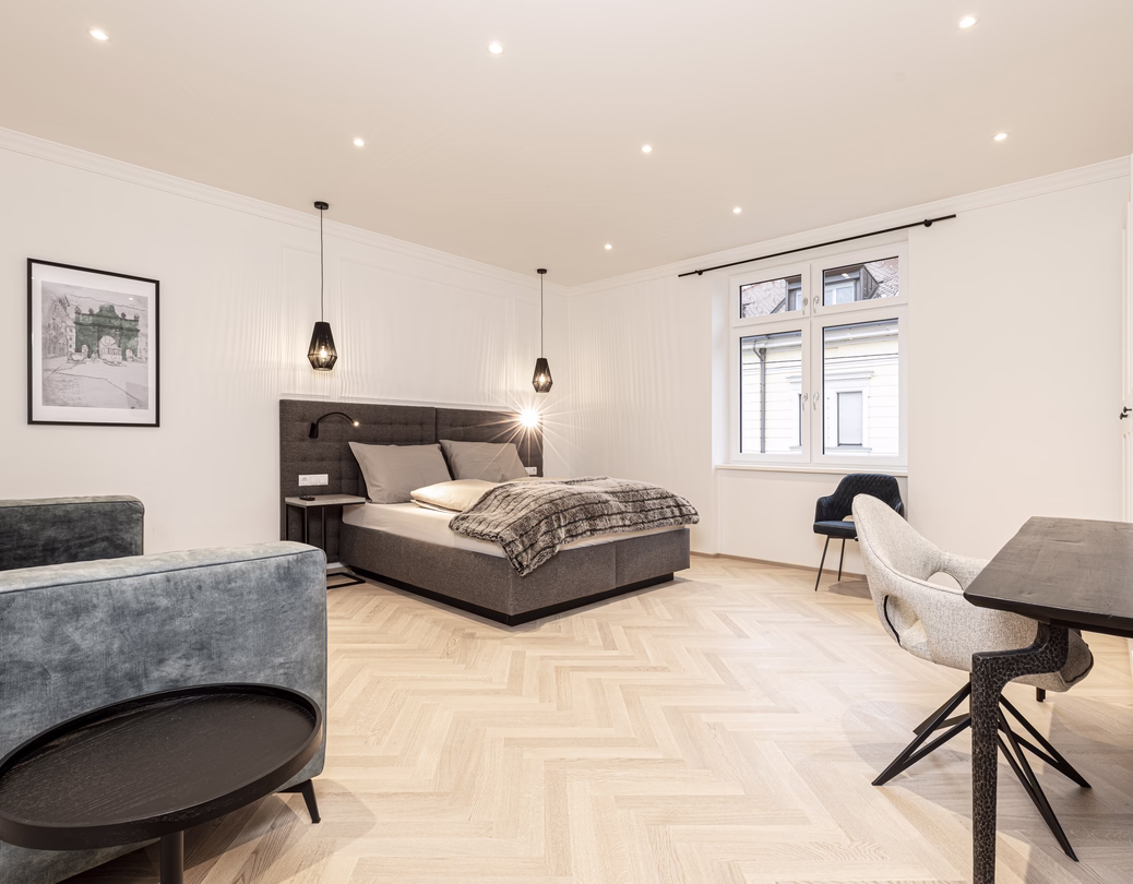 Großzügiges helles Zimmer mit Doppelbett, graugrünem Sofa, Esstisch und mehreren bequemen Polsterstühlen.