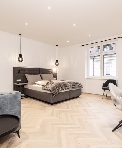 Großzügiges helles Zimmer mit Doppelbett, graugrünem Sofa, Esstisch und mehreren bequemen Polsterstühlen.