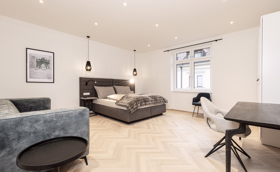 Großzügiges helles Zimmer mit Doppelbett, graugrünem Sofa, Esstisch und mehreren bequemen Polsterstühlen.