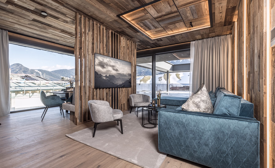 Gemütlicher Wohnbereich mit blauem Sofa, Fernseher an einer Holzlamellenwand und großen Fenstern zur winterlichen Berglandschaft.