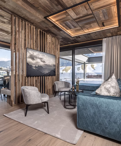 Gemütlicher Wohnbereich mit blauem Sofa, Fernseher an einer Holzlamellenwand und großen Fenstern zur winterlichen Berglandschaft.