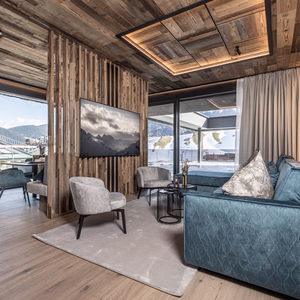 Gemütlicher Wohnbereich mit blauem Sofa, Fernseher an einer Holzlamellenwand und großen Fenstern zur winterlichen Berglandschaft.
