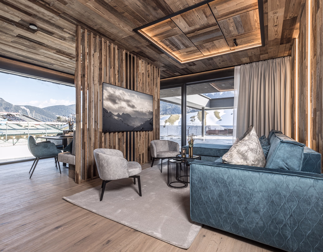 Gemütlicher Wohnbereich mit blauem Sofa, Fernseher an einer Holzlamellenwand und großen Fenstern zur winterlichen Berglandschaft.