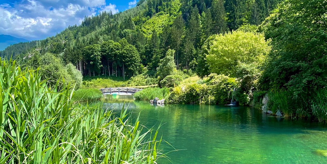 Idyllischer Badesee Aufenfeld mit klarem türkisgrünem Wasser, Schilf, kleiner Holzbrücke und bewaldeten Berghängen.