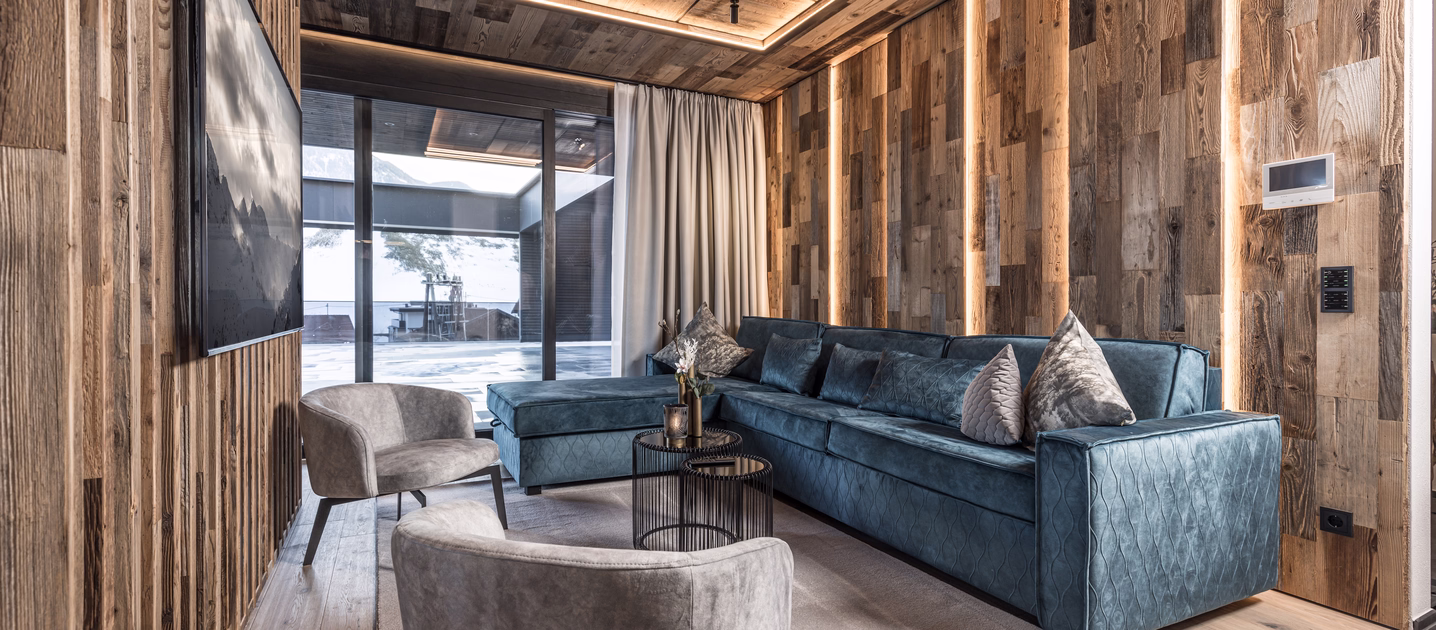 Großzügiges Wohnzimmer mit blauem Ecksofa, zwei Sesseln, Fernseher und stimmungsvoll beleuchteter Holzvertäfelung.