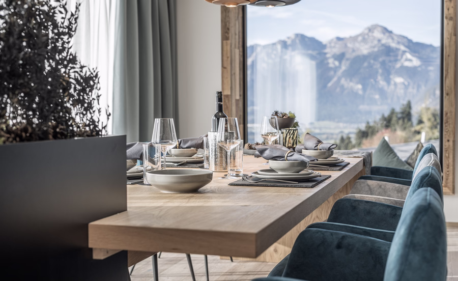 Gedeckter Esstisch mit blauen Polsterstühlen und großer Fensterfront, die einen eindrucksvollen Blick auf die Zillertaler Berge eröffnet.