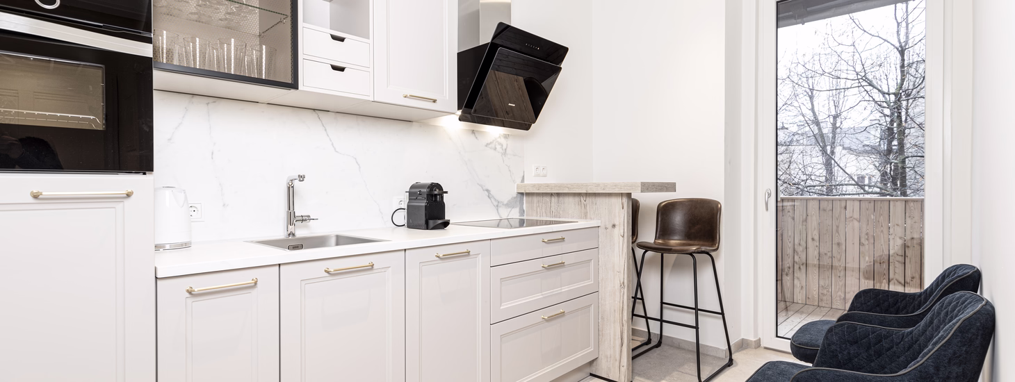 Moderne weiße Küche mit Marmoroptik, kleiner Frühstückstheke, gepolsterten Stühlen und direktem Balkonausgang.