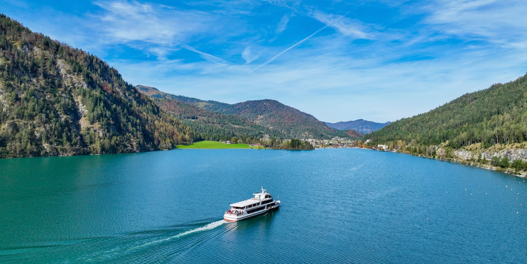 Ein Ausflugsschiff fährt über den Achensee, umgeben von bewaldeten Bergen und einer sonnigen Uferlandschaft.