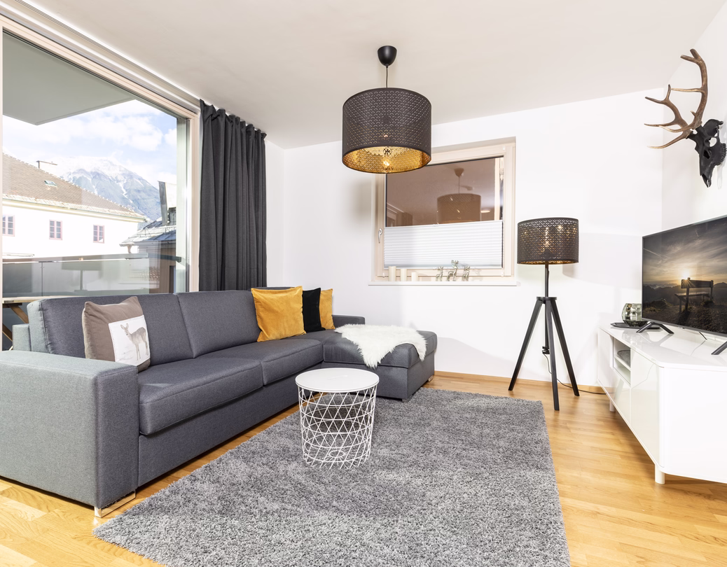 Helles Wohnzimmer mit grauem Ecksofa, Fernseher, schwarzen Leuchten und großer Balkontür mit Bergblick.