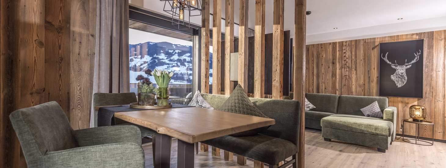Offener Wohn- und Essbereich mit Holztisch, grünen Polstermöbeln, rustikalen Holzelementen und Blick auf die verschneite Landschaft.